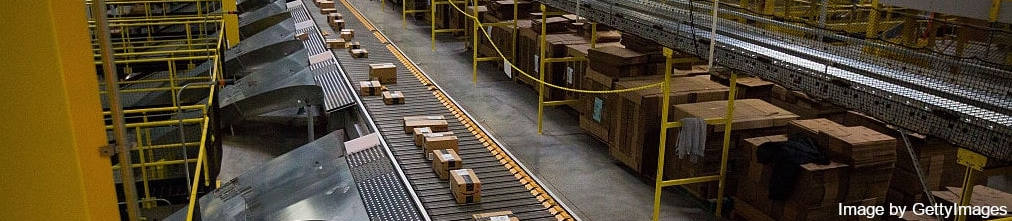Long conveyor belt in Amazon fulfillment center moving boxes with Amazon Prime stickers to another storage
