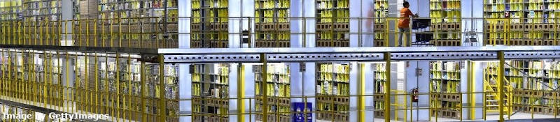 Amazon employee rolling a trolly with boxes past endless shelves of Amazon fulfillment center